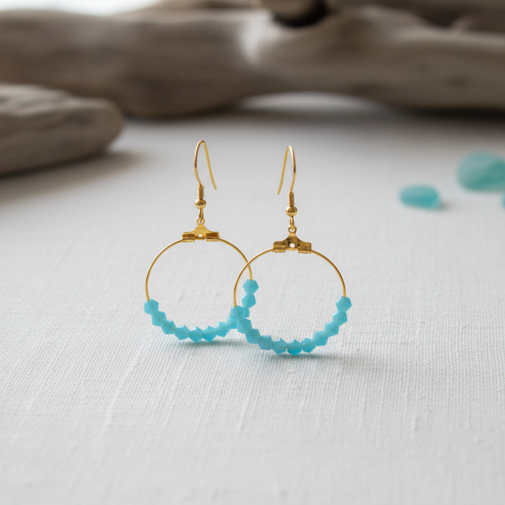 A pair of gold hoop earrings with small faceted light blue beads along the bottom curve, displayed against a plain light gray background.