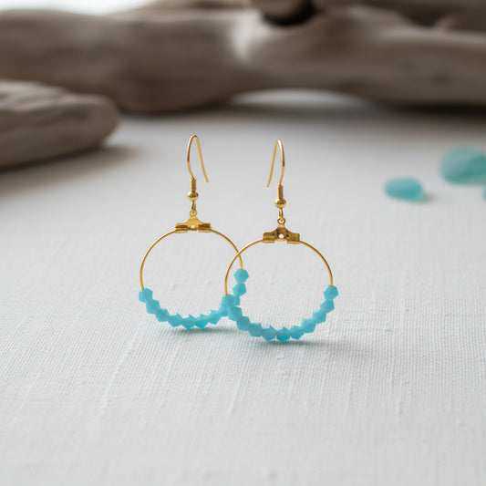A pair of gold hoop earrings with small faceted light blue beads along the bottom curve, displayed against a plain light gray background.