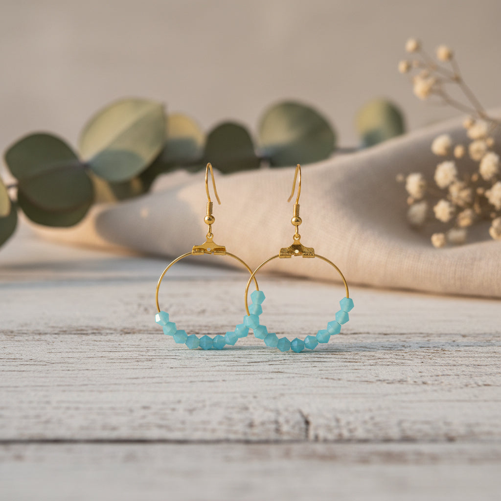 A pair of gold hoop earrings with small faceted light blue beads along the lower half, displayed against a plain light gray background.