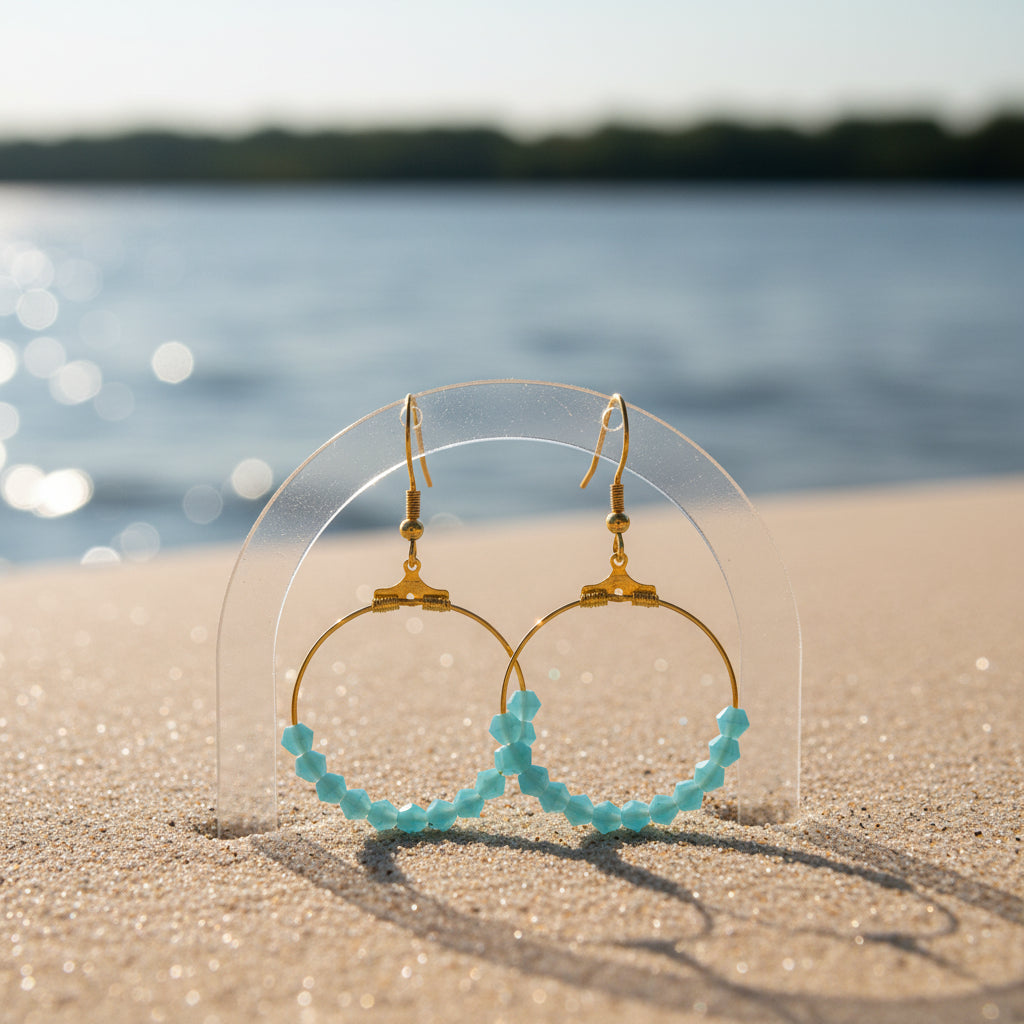 A pair of gold hoop earrings with small faceted light blue beads along the bottom curve, displayed against a plain light gray background.