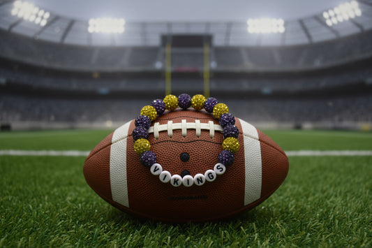 A beaded stretch bracelet featuring purple and gold glittery round beads alternating with white letter beads that spell 'VIKINGS' on a plain light gray background.