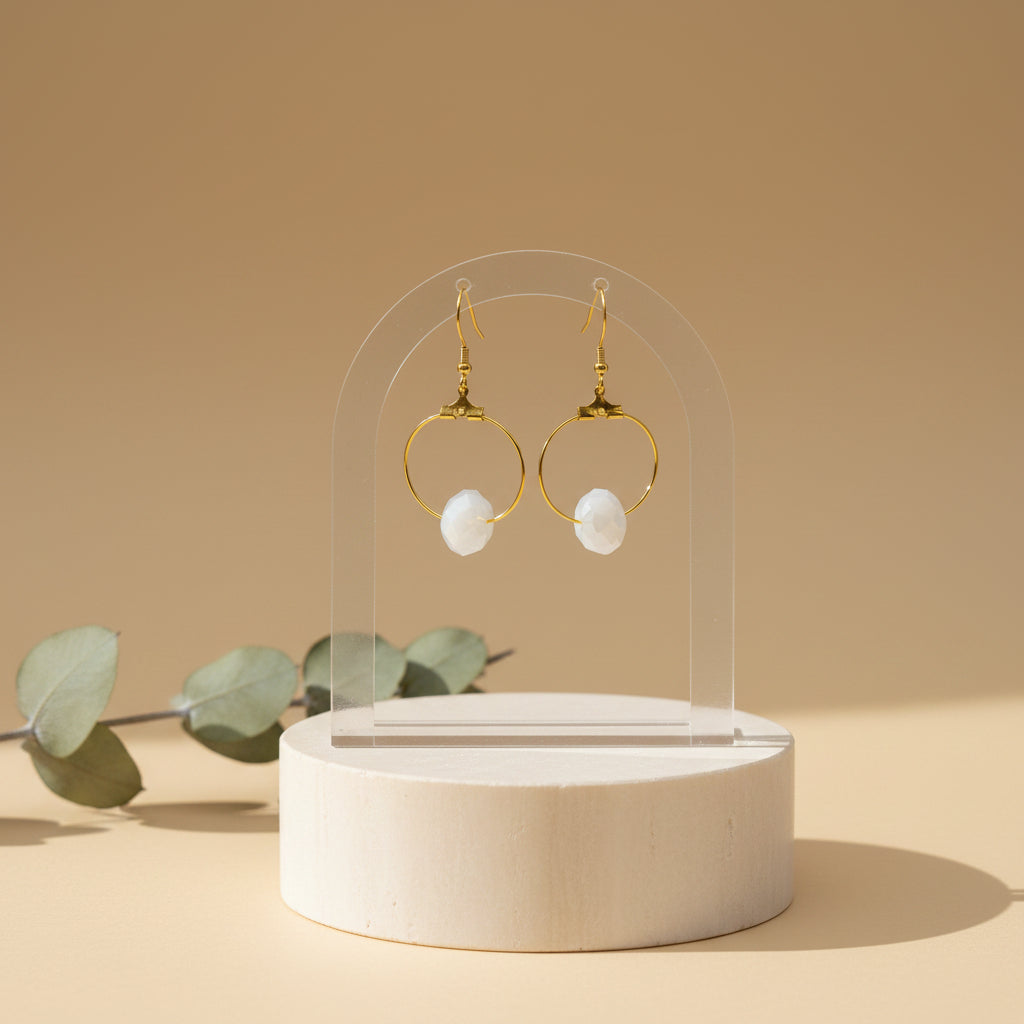 A pair of gold hoop earrings with white stone accents, displayed on a clear acrylic stand against a neutral gray background.