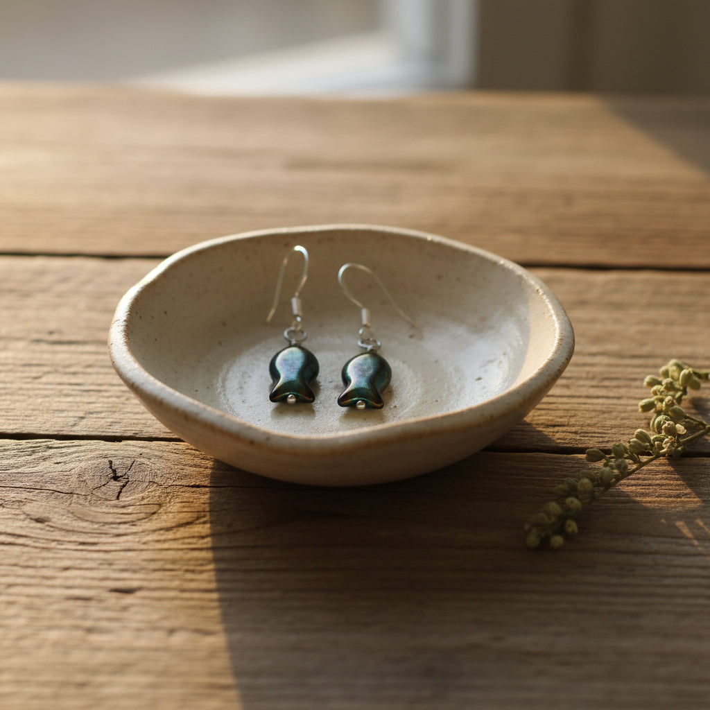 A pair of handmade fish-shaped dangle earrings with iridescent dark green and blue glass bodies and silver fishhook wires, displayed on a clear plastic stand against a neutral gray background.