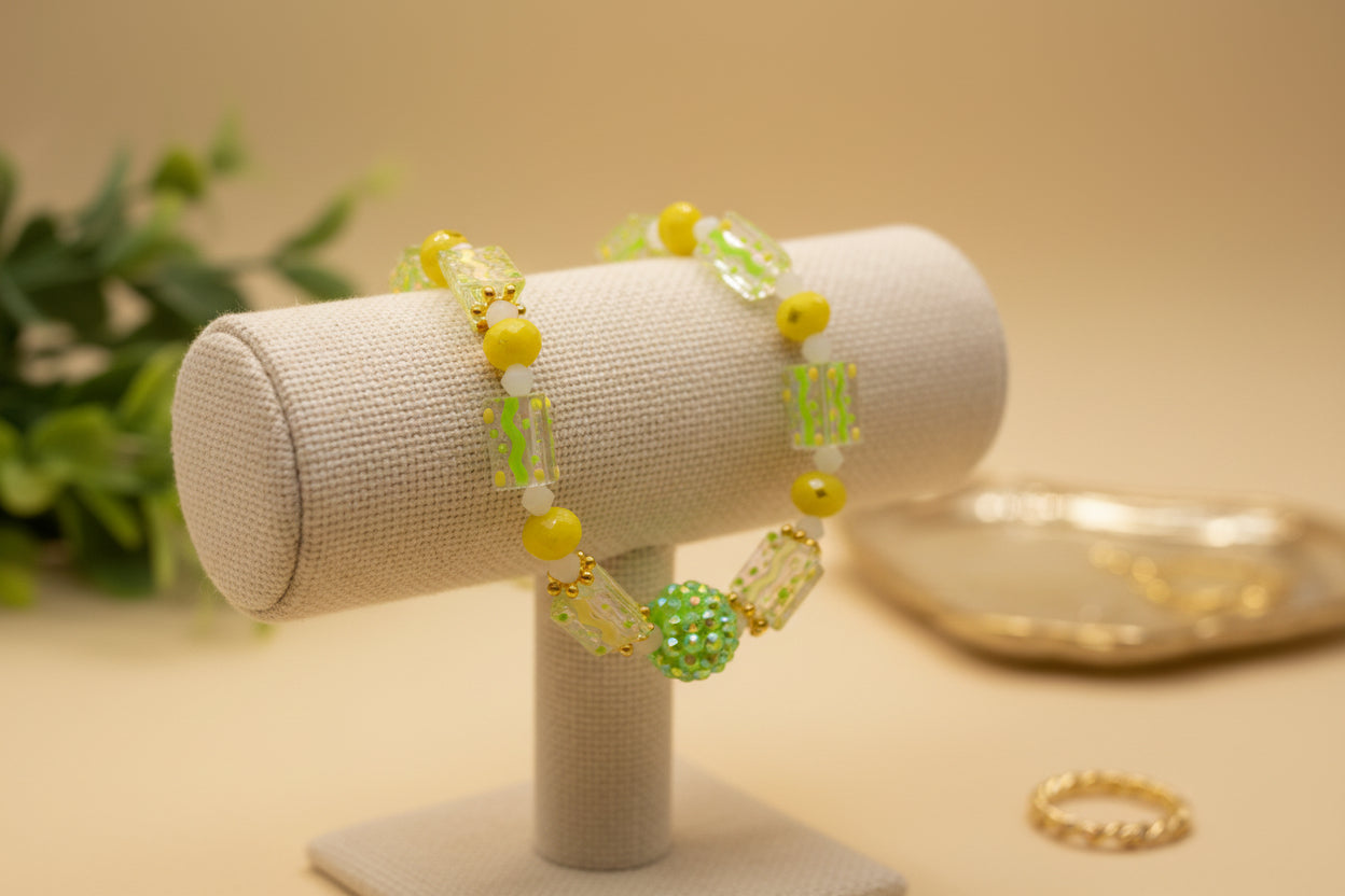 A beaded stretch bracelet featuring yellow beads, clear rectangular beads with green and yellow speckles, and a central green glittery ball accent, displayed on a neutral light gray background.