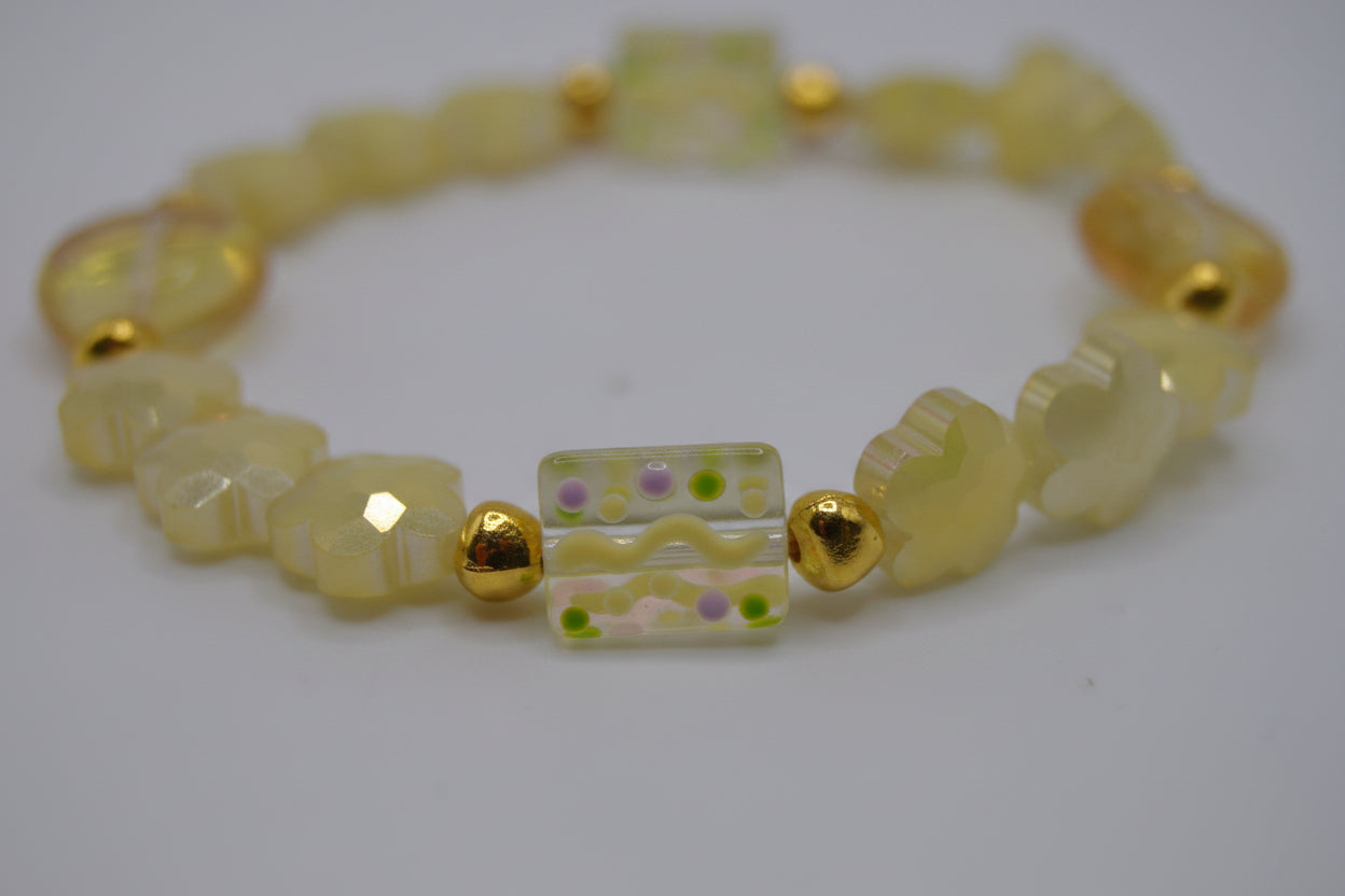 A beaded stretch bracelet featuring pale yellow translucent beads, textured elephant-shaped beads, faceted gold spacer beads, and a central rectangular glass bead with a wavy yellow and green pattern, displayed on a neutral light gray background.