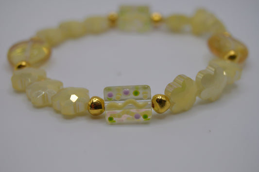 A beaded stretch bracelet featuring pale yellow translucent beads, textured elephant-shaped beads, faceted gold spacer beads, and a central rectangular glass bead with a wavy yellow and green pattern, displayed on a neutral light gray background.